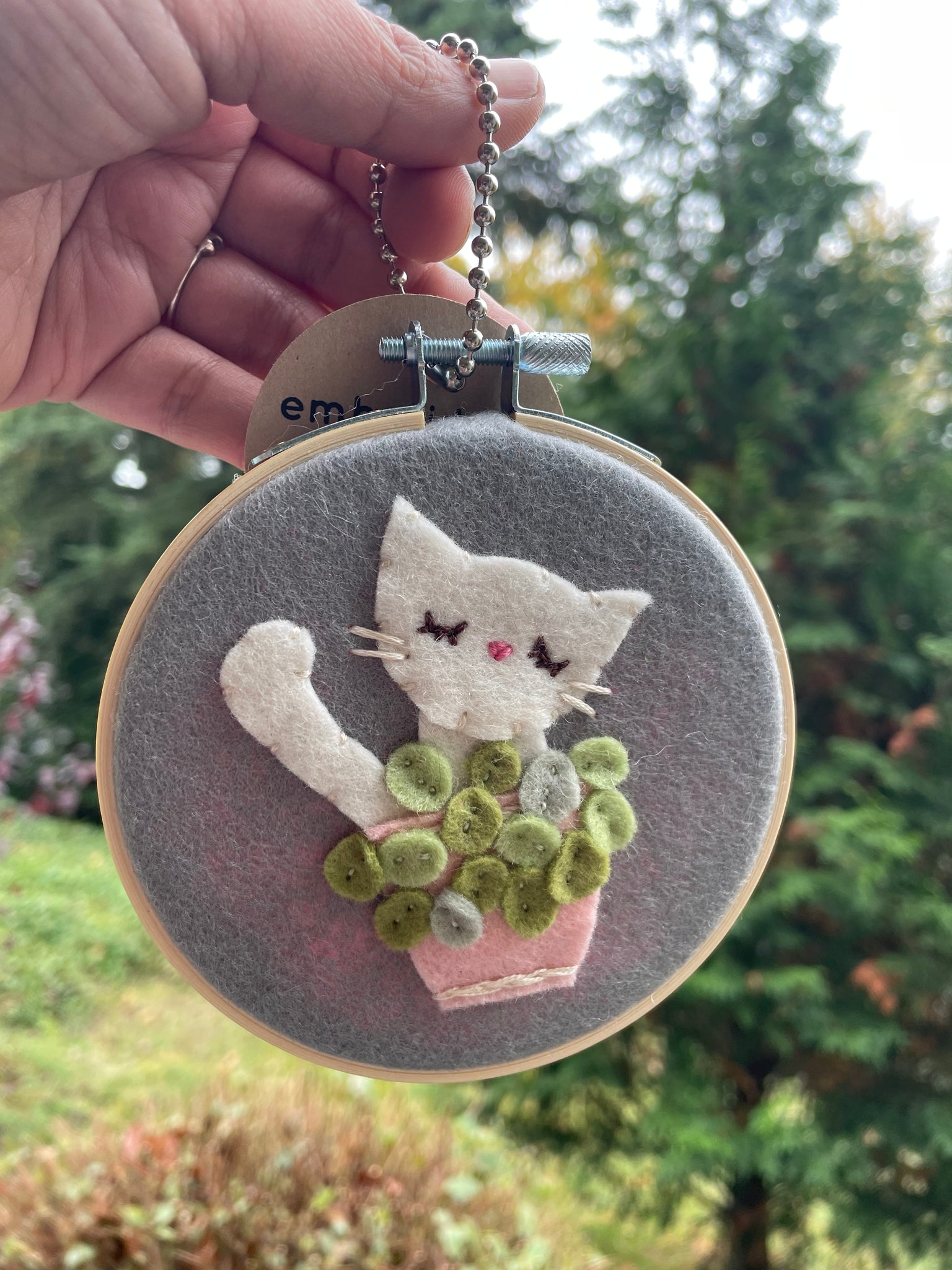 Embroidery hoop with a cat behind a potted plant.