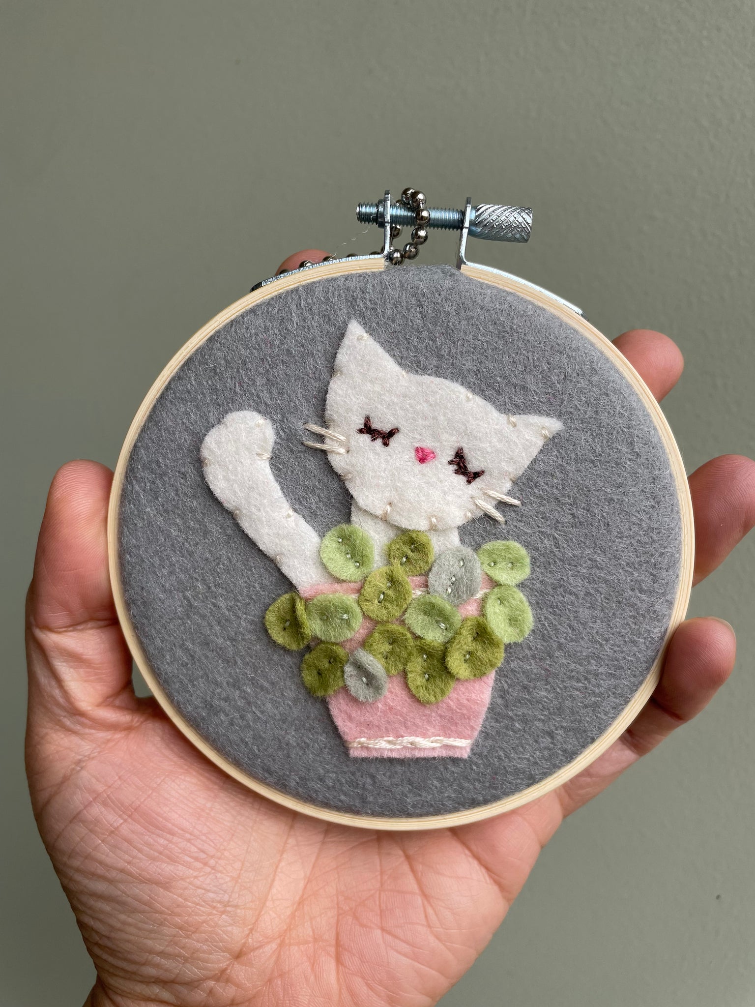 Embroidery hoop with a white cat sitting behind a potted plant held by a hand against a neutral background