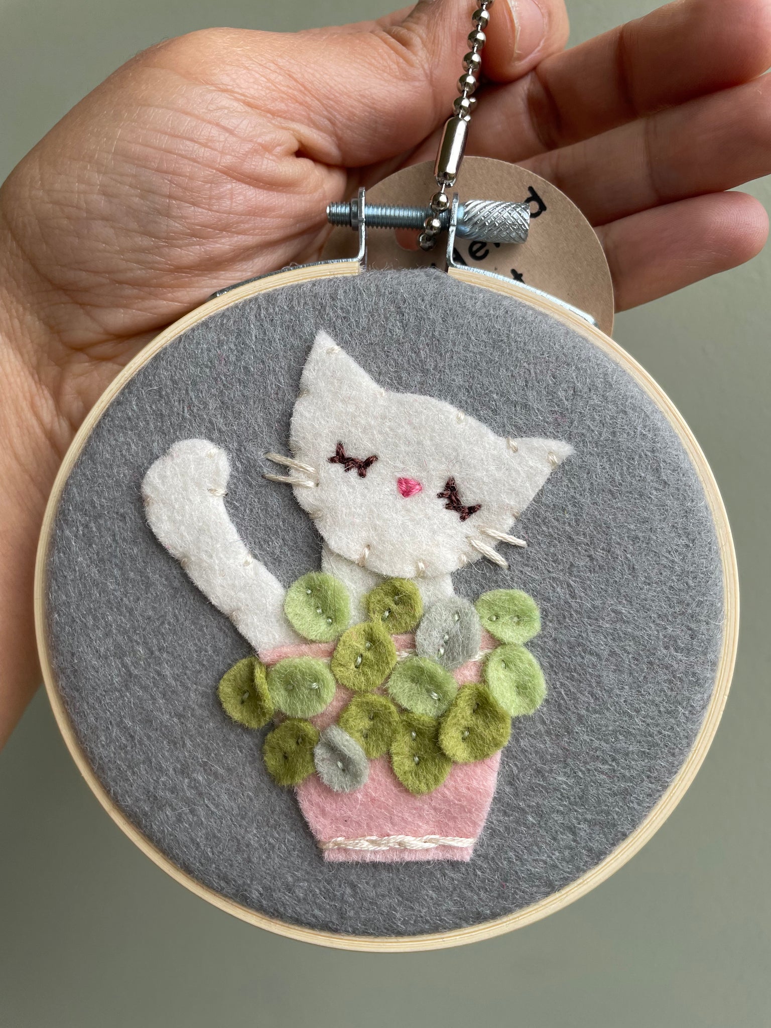 Embroidery hoop with a white cat behind a pink pot of flowers, held by a hand against a neutral background.